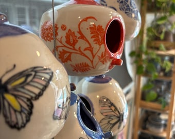 Op de afbeelding: Een verzameling hangende keramische vogelhuisjes. De vogelhuisjes zijn wit met verschillende ontwerpen, waaronder vlinders, bijen en bloempatronen. Sommige hebben rode of blauwe accenten. De vogelhuisjes zijn opgehangen aan dunne draden.
