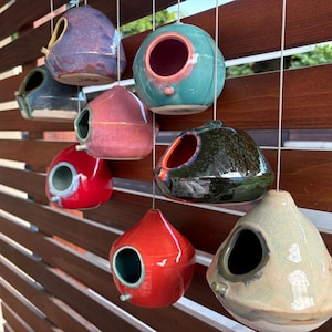 May include: A collection of eight ceramic birdhouses in various colors including red, blue, pink, green, and beige, each featuring a circular entrance hole and a small perch, suspended by white strings in a staggered arrangement.