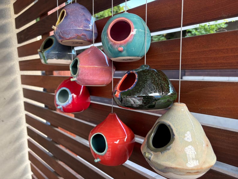 May include: A collection of eight ceramic birdhouses in various colors including red, blue, pink, green, and beige, each featuring a circular entrance hole and a small perch, suspended by white strings in a staggered arrangement.