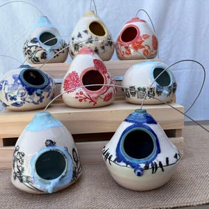 May include: A collection of eight ceramic birdhouses with various hand-painted designs, including floral patterns, owls, and birds. Each teardrop-shaped house features a circular entrance hole and a metal wire loop for hanging, displayed on a wooden stand.