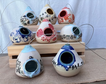 May include: A collection of eight ceramic birdhouses with various hand-painted designs, including floral patterns, owls, and birds. Each teardrop-shaped house features a circular entrance hole and a metal wire loop for hanging, displayed on a wooden stand.