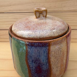 May include: A cylindrical ceramic jar with a matching lid featuring a knob handle. The exterior displays vertical stripes of green, blue, purple, and orange glaze over a textured clay body. The lid has a speckled cream and tan finish.