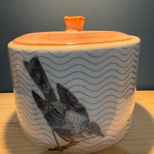 May include: A cylindrical ceramic jar with a lid featuring a bright orange top and knob. The body is white with a repeating pattern of thin blue wavy lines and a detailed black and white illustration of a small bird perched on a branch.