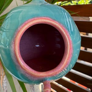 May include: A spherical ceramic birdhouse with a glossy teal exterior and a smooth, rounded opening. The rim of the entrance is highlighted with a vibrant coral-pink glaze that transitions into the deep pink interior of the hollow vessel.