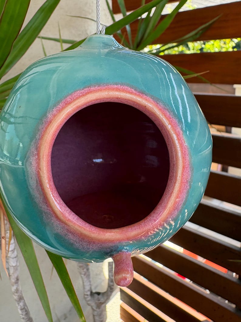 May include: A spherical ceramic birdhouse with a glossy teal exterior and a smooth, rounded opening. The rim of the entrance is highlighted with a vibrant coral-pink glaze that transitions into the deep pink interior of the hollow vessel.