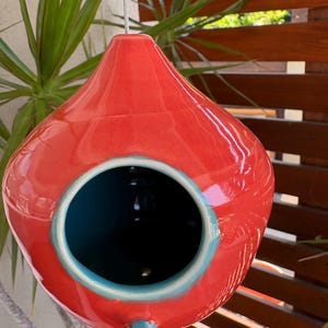 May include: A teardrop-shaped ceramic birdhouse with a glossy red exterior and a contrasting teal interior. The structure features a circular opening on the front, a small teal perch below the entrance, and a metal wire for hanging.