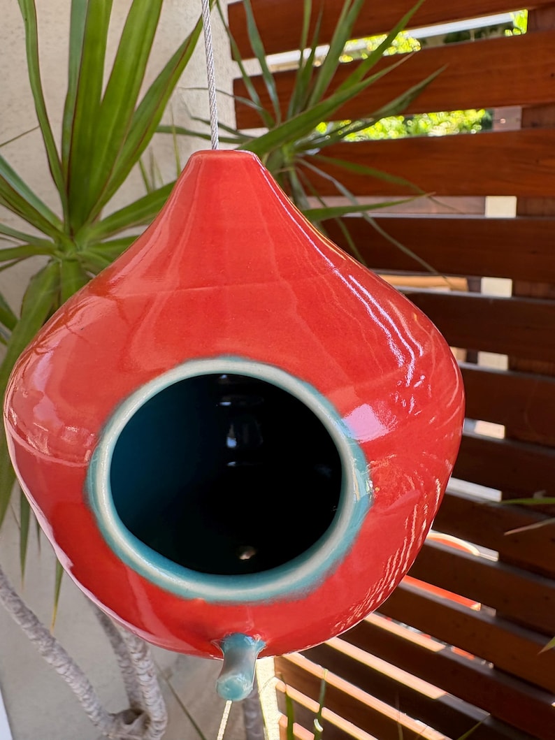 May include: A teardrop-shaped ceramic birdhouse with a glossy red exterior and a contrasting teal interior. The structure features a circular opening on the front, a small teal perch below the entrance, and a metal wire for hanging.