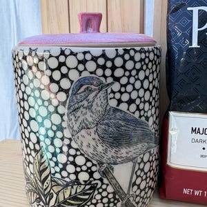 May include: A cylindrical ceramic canister with a pink lid and a black and white polka dot pattern. The front features a detailed illustration of a bird perched on a branch with leaves. The canister is positioned next to a dark coffee bag.