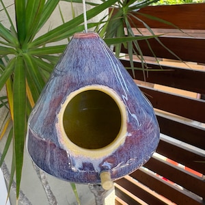 May include: A teardrop-shaped ceramic birdhouse with a glossy glaze featuring blended shades of deep purple, blue, and hints of red. It has a large circular opening with a yellow interior and a small wooden perch extending from the bottom.