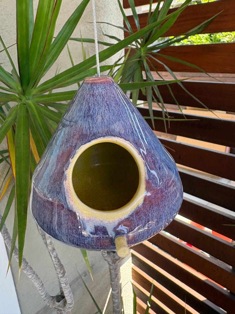May include: A teardrop-shaped ceramic birdhouse with a glossy glaze featuring blended shades of deep purple, blue, and hints of red. It has a large circular opening with a yellow interior and a small wooden perch extending from the bottom.