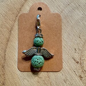 May include: A charm featuring a silver angel design with green, textured beads. The angel has silver wings and a decorative top. The charm is attached to a silver clasp and a brown tag.