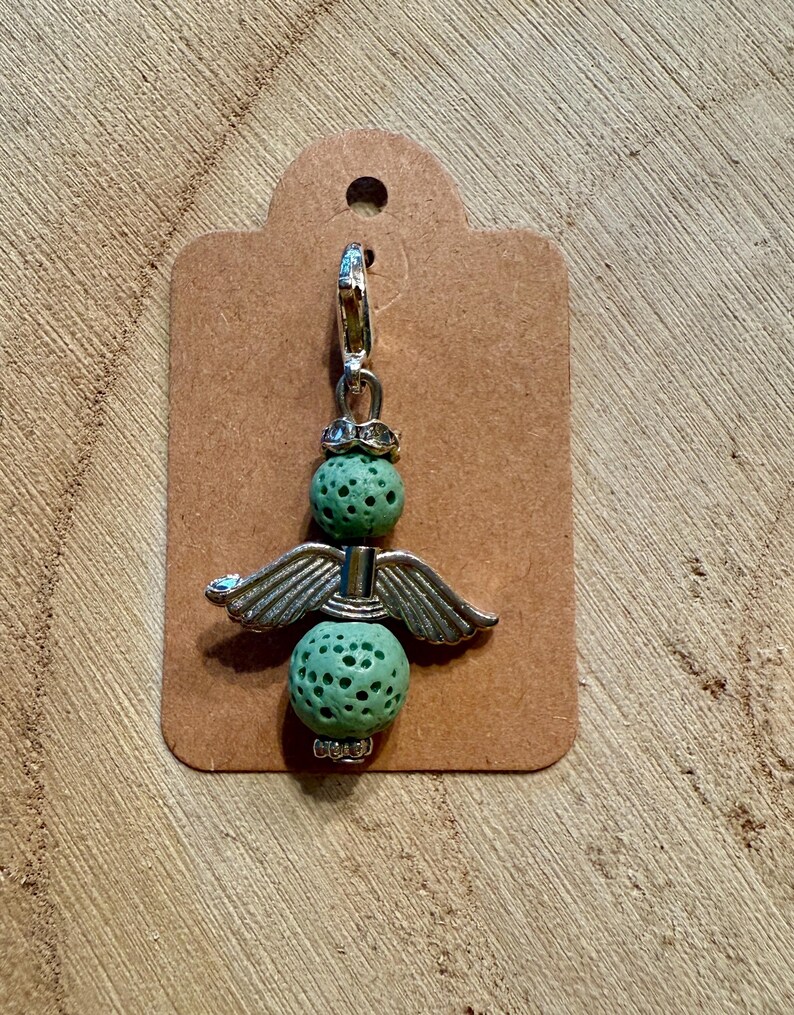 May include: A charm featuring a silver angel design with green, textured beads. The angel has silver wings and a decorative top. The charm is attached to a silver clasp and a brown tag.
