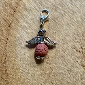 May include: A silver-toned angel charm with a lobster clasp. The charm features a pink, porous lava bead body, silver wings, and a decorative head. The charm is designed to be attached to a bracelet or necklace.