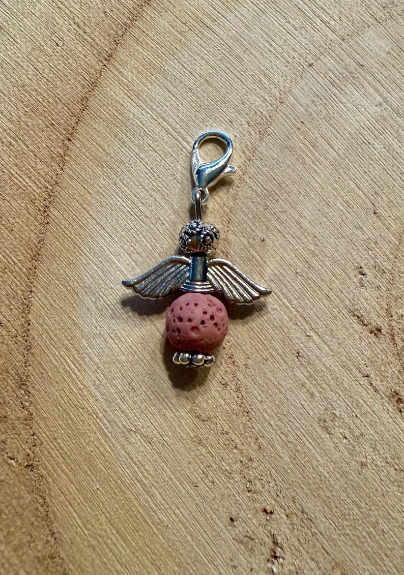 May include: A silver-toned angel charm with a lobster clasp. The charm features a pink, porous lava bead body, silver wings, and a decorative head. The charm is designed to be attached to a bracelet or necklace.