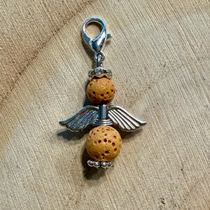 May include: A silver-toned charm in the shape of an angel, featuring two textured, light brown spheres and silver wings. The charm has a lobster clasp for easy attachment. The angel charm is a decorative accessory.