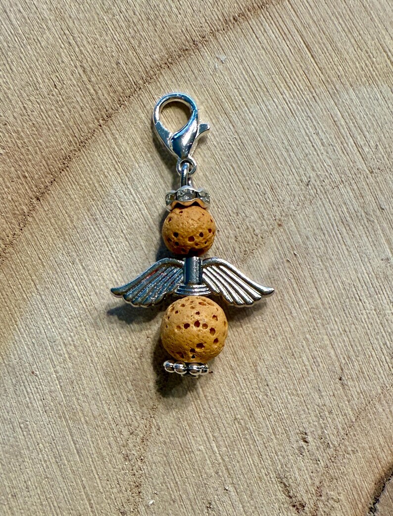 May include: A silver-toned charm in the shape of an angel, featuring two textured, light brown spheres and silver wings. The charm has a lobster clasp for easy attachment. The angel charm is a decorative accessory.