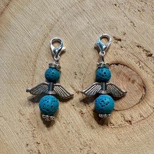 May include: Two angel-themed charms with silver-colored metal wings and clasps. Each charm features two turquoise-colored, porous, spherical beads. The charms are displayed on a wooden surface.
