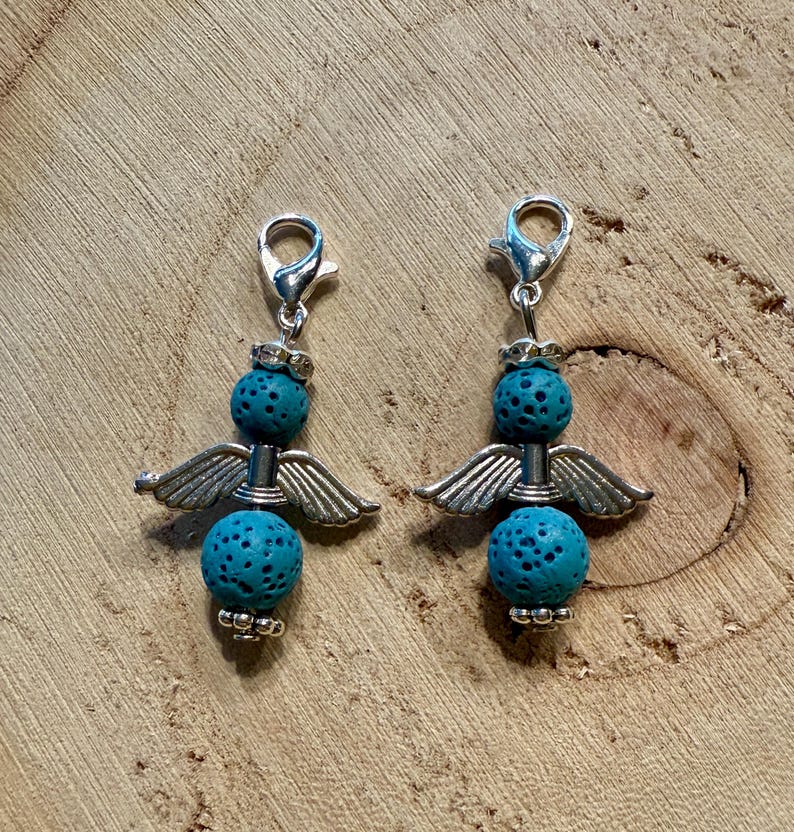 May include: Two angel-themed charms with silver-colored metal wings and clasps. Each charm features two turquoise-colored, porous, spherical beads. The charms are displayed on a wooden surface.