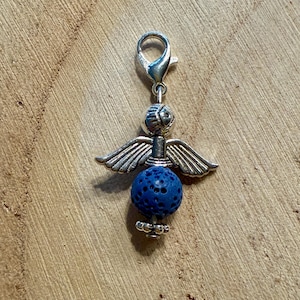 May include: A silver-toned angel charm with outstretched wings and a blue lava bead body. The charm has a lobster clasp for easy attachment to a bracelet or necklace. The angel's head is detailed with etched lines.