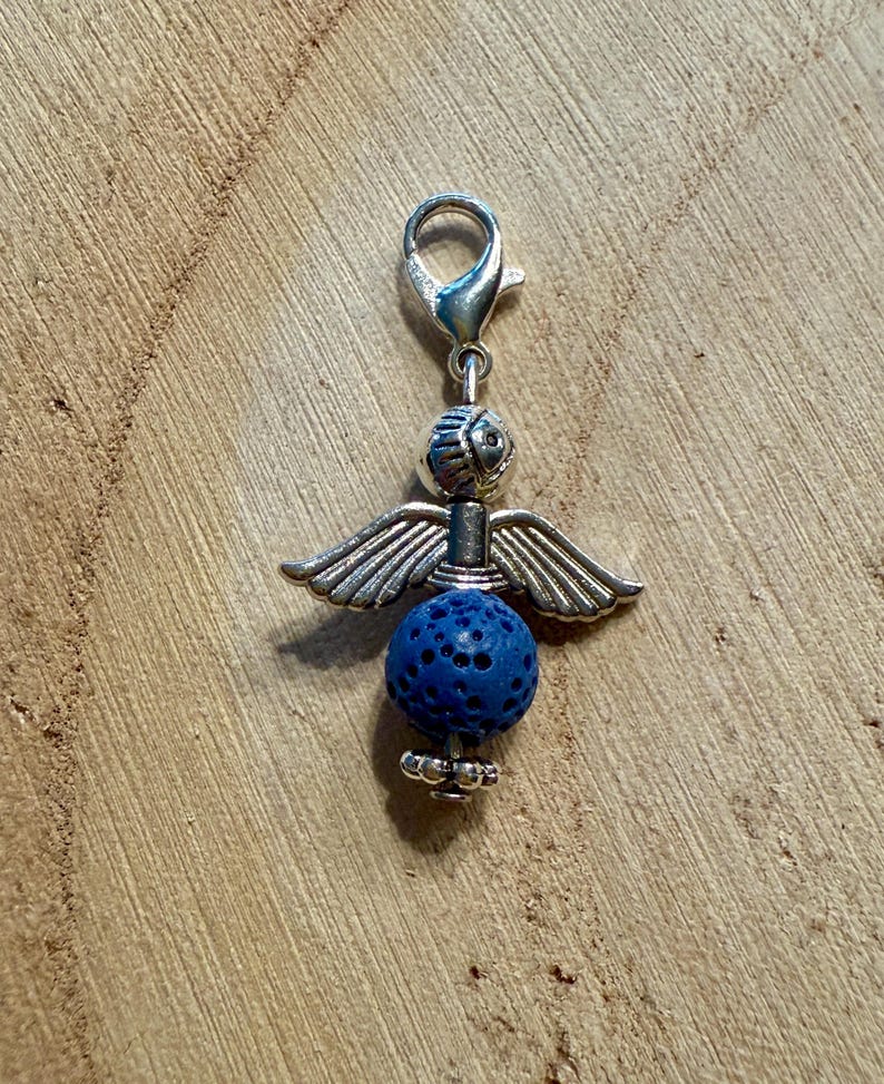 May include: A silver-toned angel charm with outstretched wings and a blue lava bead body. The charm has a lobster clasp for easy attachment to a bracelet or necklace. The angel's head is detailed with etched lines.