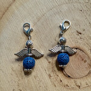May include: Two silver-toned angel charms with blue lava rock beads. Each charm features a detailed angel design with outstretched wings and a small, round blue bead. The charms are displayed on a wooden surface.