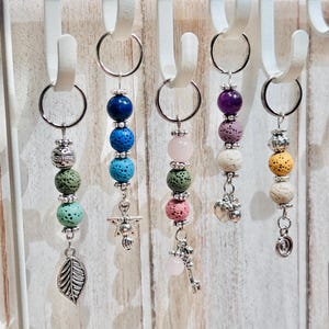 Könnte beinhalten: Eine Sammlung von Schlüsselanhängern mit Perlen in verschiedenen Farben, darunter Blau, Rosa, Grün, Lila und Gelb. Jeder Schlüsselanhänger hat einen silberfarbenen Anhänger, wie ein Blatt, einen Engel oder ein Herz. Die Schlüsselanhänger hängen an weißen Haken.