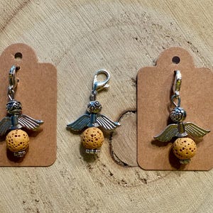 May include: Three charm pendants with angel wing designs. Each charm features a silver-colored metal top with wings, a textured orange-yellow bead, and a silver clasp. The charms are displayed on brown tags against a wooden background.