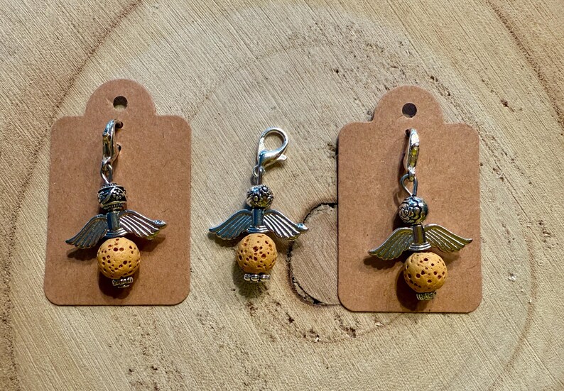 May include: Three charm pendants with angel wing designs. Each charm features a silver-colored metal top with wings, a textured orange-yellow bead, and a silver clasp. The charms are displayed on brown tags against a wooden background.