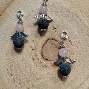 May include: Three angel charms with silver-colored wings and clasps. Each charm features a pink gemstone head, a dark gray lava rock body, and silver-colored metal accents. The charms are displayed on a wooden surface.
