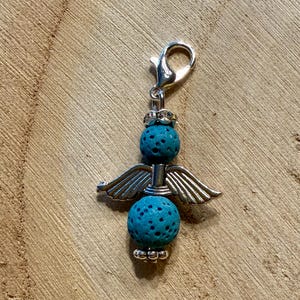 May include: A silver-toned angel charm with turquoise porous beads and detailed wings. The charm has a lobster clasp for easy attachment. The angel's wings are spread, and the charm is set against a wooden background.