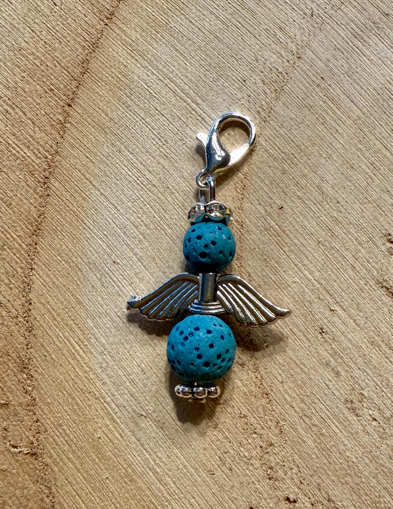 May include: A silver-toned angel charm with turquoise porous beads and detailed wings. The charm has a lobster clasp for easy attachment. The angel's wings are spread, and the charm is set against a wooden background.