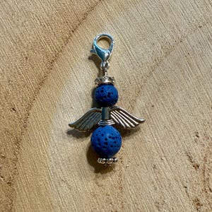May include: A silver-toned charm featuring an angel design. The charm has two blue lava stone spheres, silver wings, and a clasp. The charm is displayed on a wooden surface.