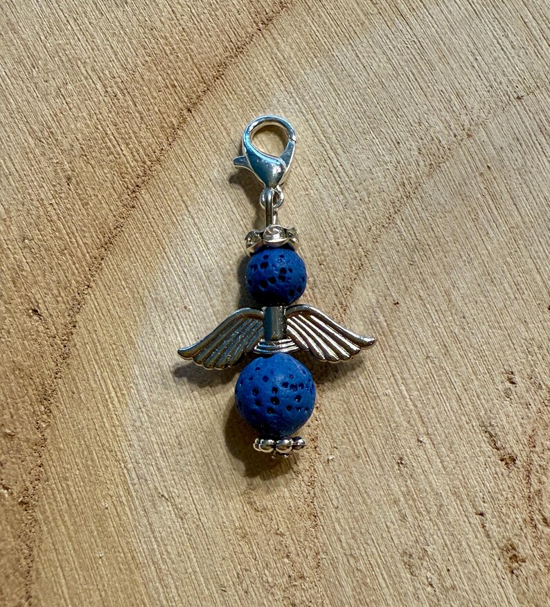 May include: A silver-toned charm featuring an angel design. The charm has two blue lava stone spheres, silver wings, and a clasp. The charm is displayed on a wooden surface.