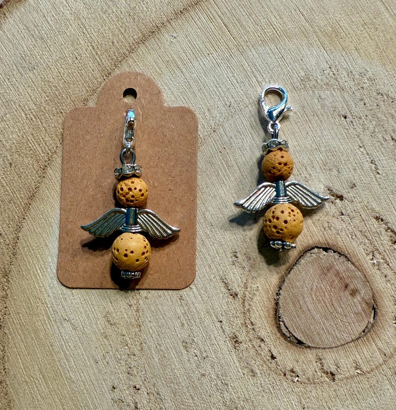 May include: Two angel charm pendants. Each charm features two textured, light brown spherical beads, silver-colored wings, and a silver-colored clasp. One charm is attached to a brown tag.