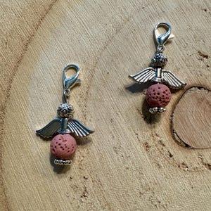 May include: Two silver-toned angel charms with pink lava rock beads. Each charm features a detailed angel design with wings and a lobster clasp for easy attachment. The charms are displayed on a wooden surface.