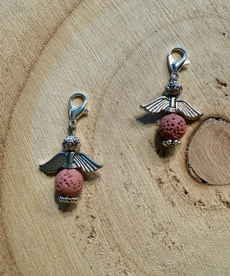 May include: Two silver-toned angel charms with pink lava rock beads. Each charm features a detailed angel design with wings and a lobster clasp for easy attachment. The charms are displayed on a wooden surface.