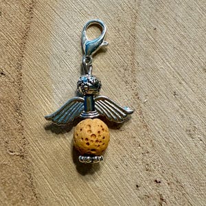 May include: A silver-colored angel charm with a lobster clasp. The charm features a textured, spherical, light brown lava bead body, silver wings, and a decorative head. The charm is designed to be attached to a bracelet or necklace.