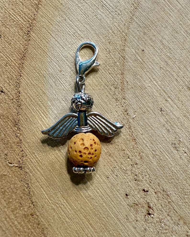 May include: A silver-colored angel charm with a lobster clasp. The charm features a textured, spherical, light brown lava bead body, silver wings, and a decorative head. The charm is designed to be attached to a bracelet or necklace.