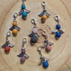 Könnte beinhalten: Eine Sammlung von Engeln-Anhängern mit silberfarbenen Flügeln und Verschlüssen. Jeder Anhänger hat eine andersfarbige Perle, darunter blau, orange, rosa und grau. Die Anhänger werden auf einer Holzoberfläche präsentiert.