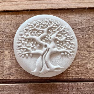 May include: A round, off-white decorative piece featuring a detailed relief of a tree with leaves and fruit. The tree design is centered on the circular object. The piece is set against a wooden background.