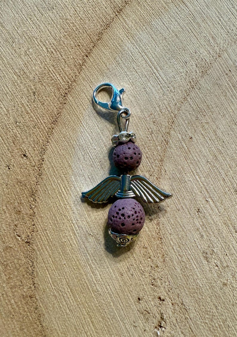 May include: A silver-toned charm with a lobster clasp, featuring two textured purple spheres and silver wings. The charm has a decorative design at the top and bottom, creating an angel-like appearance. Ideal for jewelry making.