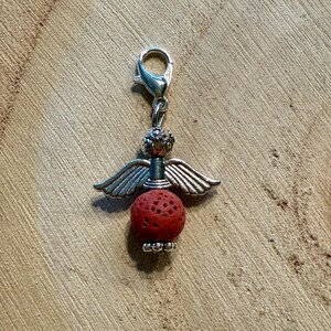 May include: A silver-colored angel charm with a red, textured bead body and silver wings. The charm has a lobster clasp for easy attachment to a bracelet or necklace. The charm is on a wooden surface.