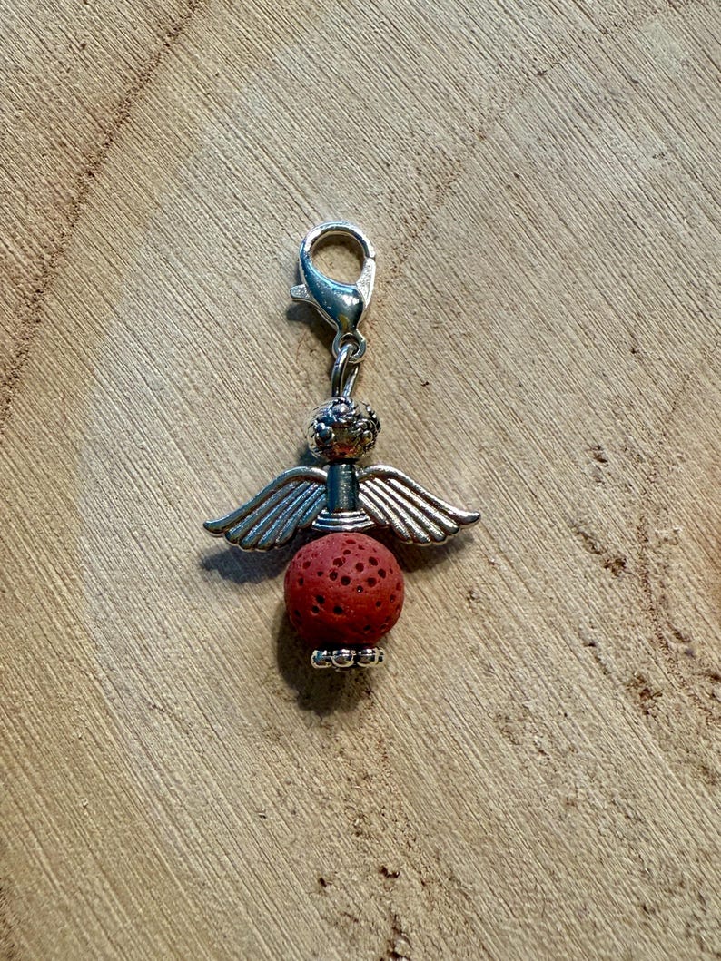 May include: A silver-colored angel charm with a red, textured bead body and silver wings. The charm has a lobster clasp for easy attachment to a bracelet or necklace. The charm is on a wooden surface.