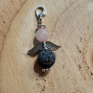 May include: A charm featuring an angel design. The charm has a silver-colored clasp and a pink bead for the head. Below the head are silver-colored wings, and a dark gray, textured bead forms the body. The charm is on a wooden surface.
