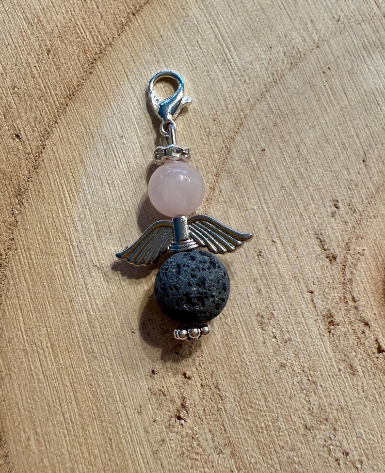May include: A charm featuring an angel design. The charm has a silver-colored clasp and a pink bead for the head. Below the head are silver-colored wings, and a dark gray, textured bead forms the body. The charm is on a wooden surface.
