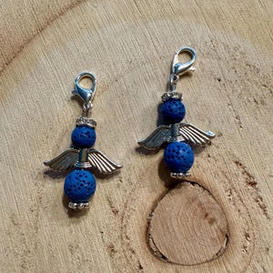 May include: Two angel-themed charms with silver-colored metal wings and clasps. Each charm features two blue, textured beads. The charms are displayed on a wooden surface.