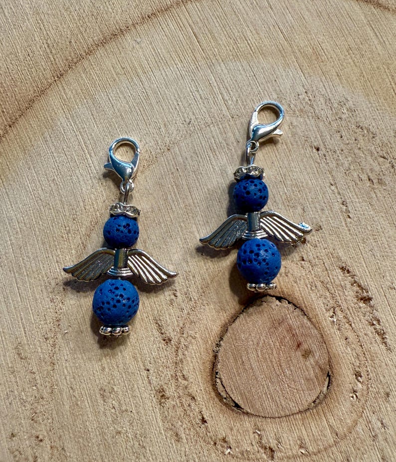 May include: Two angel-themed charms with silver-colored metal wings and clasps. Each charm features two blue, textured beads. The charms are displayed on a wooden surface.