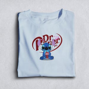 Może przedstawiać: Jasnoniebieska koszulka z postacią z kresk&oacute;wki Stitch w słuchawkach i trzymającą puszkę Dr. Pepper. Logo Dr. Pepper znajduje się nad Stitchem. Koszulka jest starannie złożona.