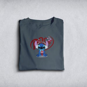 Może przedstawiać: Złożona, ciemnoszara koszulka z nadrukiem kresk&oacute;wki Stitcha w słuchawkach i trzymającego nap&oacute;j. Projekt zawiera nuty muzyczne i czerwone serce z tekstem.