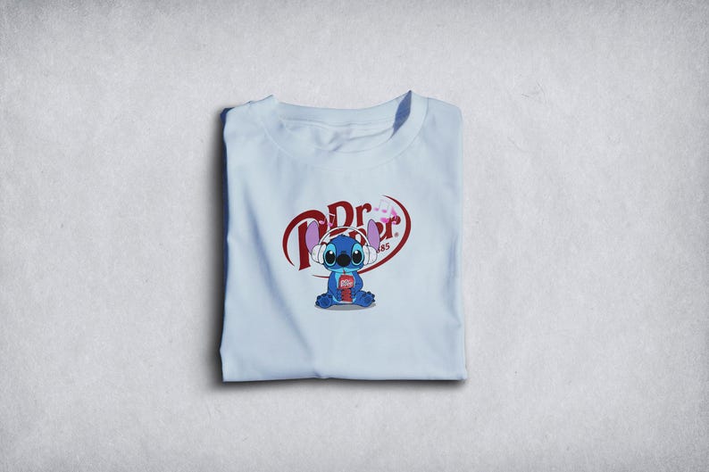 Może przedstawiać: Jasnoniebieska koszulka z postacią z kresk&oacute;wki Stitch w słuchawkach i trzymającą puszkę Dr. Pepper. Logo Dr. Pepper znajduje się nad Stitchem. Koszulka jest starannie złożona.